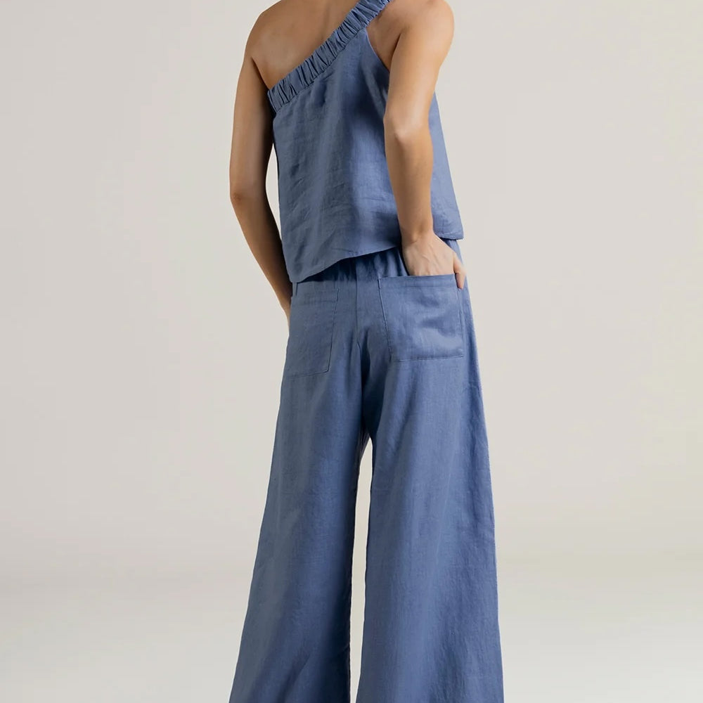 Person wearing a blue one shoulder top and wide-leg pants on a plain background.