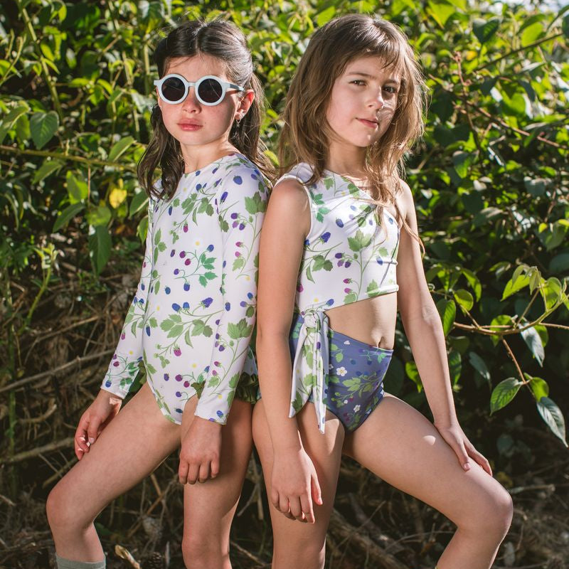 Two young girls in matching one-piece floral swimsuits and yellow boots standing in a natural setting.