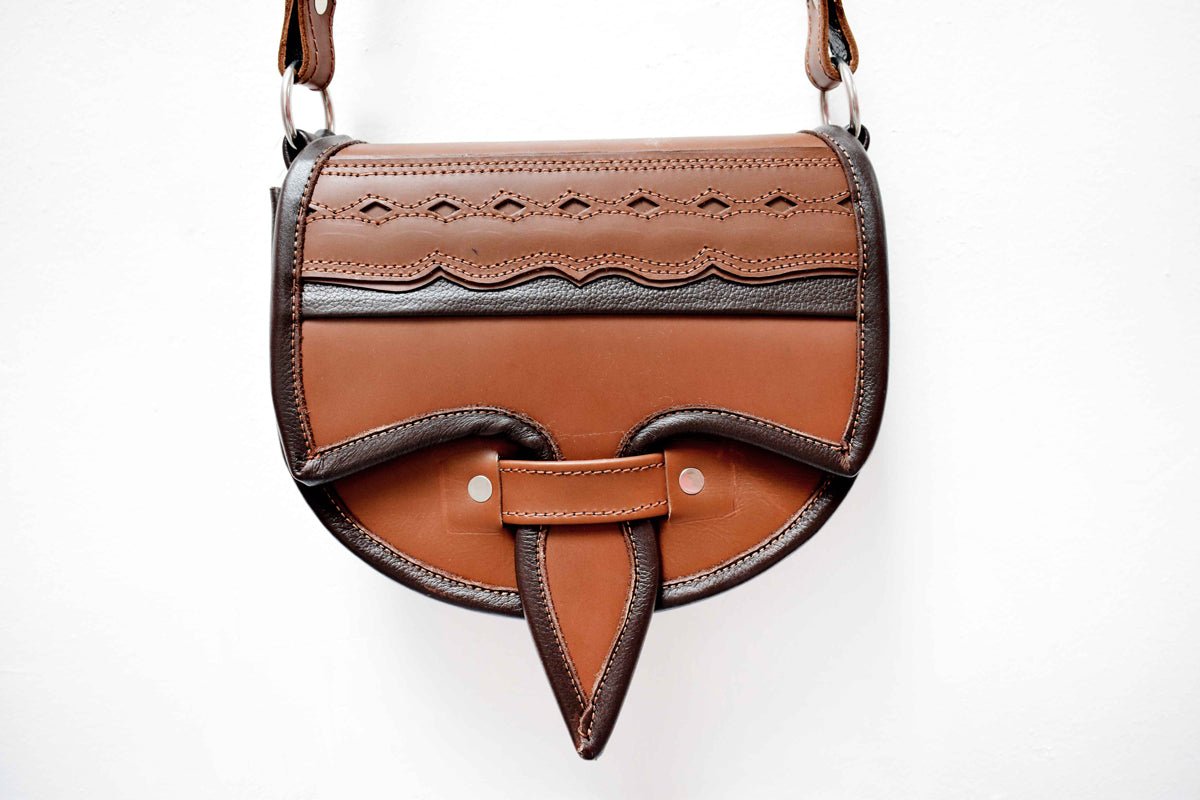 A two-toned brown leather crossbody saddle bag with multiple pockets and hardware accents, handmade in Colombia.
