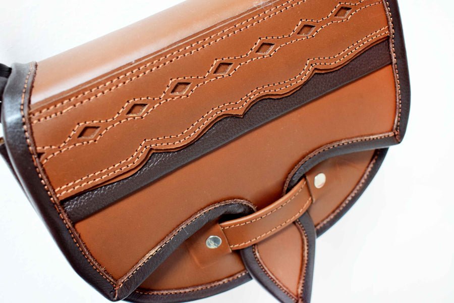 A two-toned brown leather crossbody saddle bag with multiple pockets and hardware accents, handmade in Colombia.
