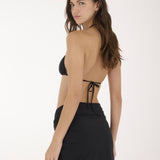 A woman modeling a black two-piece swimwear set consisting of a triangle bikini top and a high-waisted mini skirt with side ties, photographed against a plain white studio background.