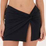 A close-up of a model wearing a black wrap-style bikini skirt with a twisted front detail and a side slit, shown against a plain light background.