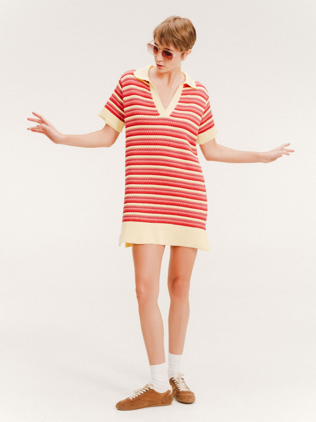 Woman wearing a short striped knit polo dress in red, coral, and pale yellow with a collared V-neck.
