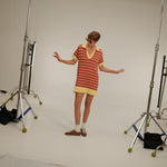 Backstage look at a model wearing a short striped knit polo dress in red, coral, and pale yellow with a collared V-neck.