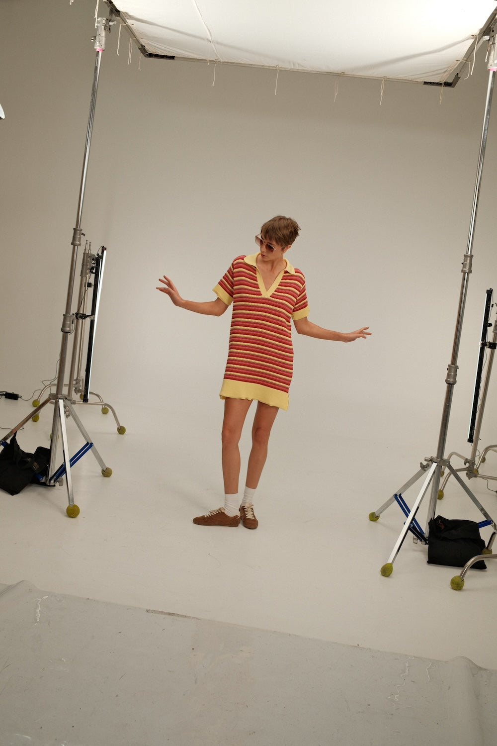 Backstage look at a model wearing a short striped knit polo dress in red, coral, and pale yellow with a collared V-neck.