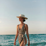 Woman walking at the beach. She's wearing a hat and a black and white one piece with a removable belt on the waist.