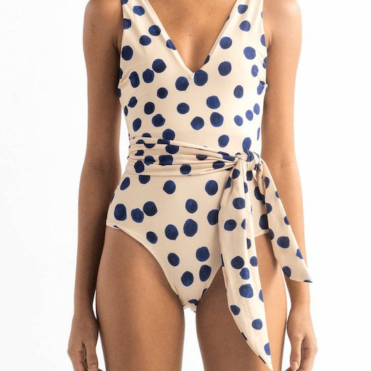 Front of a model wearing the Vero One Piece by Encantadore Swimwear. It has blue dots and a matching belt tied on the waist.