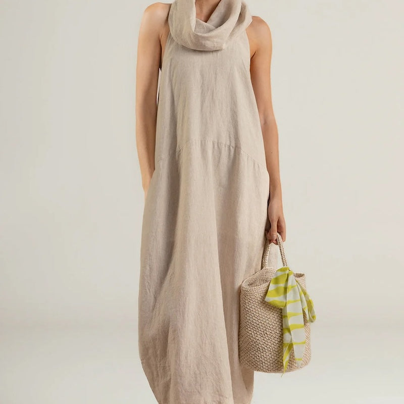 Woman wearing a beige sleeveless dress holding a woven bag on a white background.