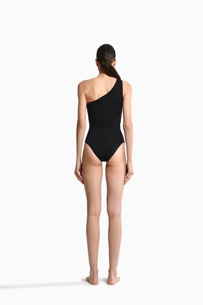  Image of the back of a woman wearing a black swimsuit with draping on the front. It's a one shoulder fit. The purpose of the image is to highlight the swimsuit because it is for sale at EVAMAIA Boutique.
