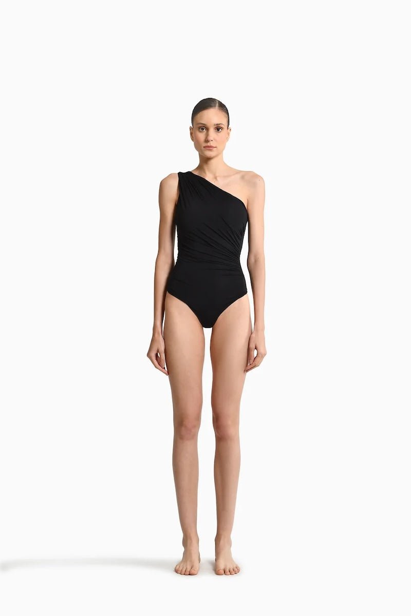 Image of a woman wearing a black swimsuit with draping on the front. It's a one shoulder fit. The purpose of the image is to highlight the swimsuit because it is for sale at EVAMAIA Boutique.