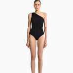 Image of a woman wearing a black swimsuit with draping on the front. It's a one shoulder fit. The purpose of the image is to highlight the swimsuit because it is for sale at EVAMAIA Boutique.