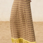 Person wearing a long, patterned skirt with yellow fringe on a sandy background.
