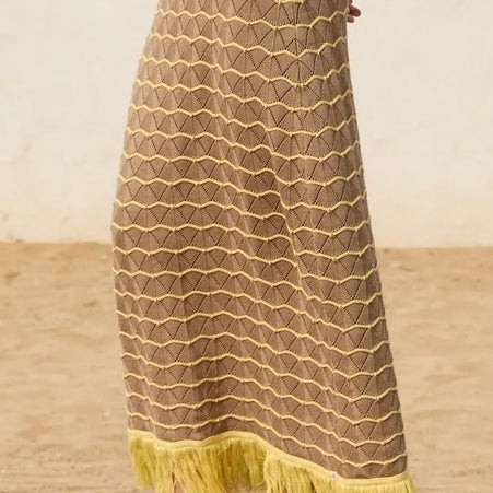 Person wearing a long, patterned skirt with yellow fringe on a sandy background.