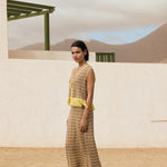 Woman in a yellow and brown dress standing in a desert-like setting with a white wall and green structure in the background.
