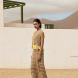 Woman in a yellow and brown dress standing in a desert-like setting with a white wall and green structure in the background.