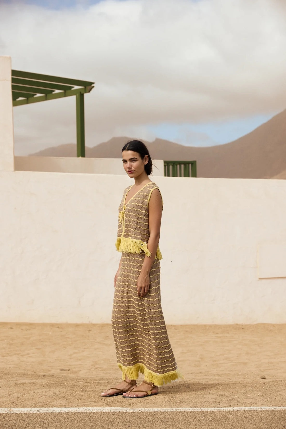 Woman in a yellow and brown dress standing in a desert-like setting with a white wall and green structure in the background.