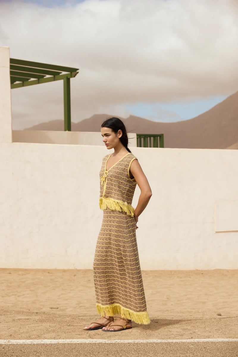 Woman in a long, patterned dress with yellow accents standing in a desert-like setting.