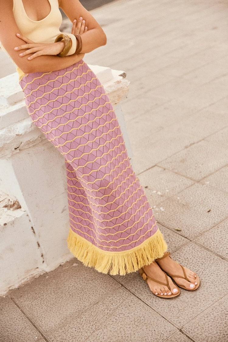 Person wearing a purple and yellow patterned skirt with tassels on a stone pavement.