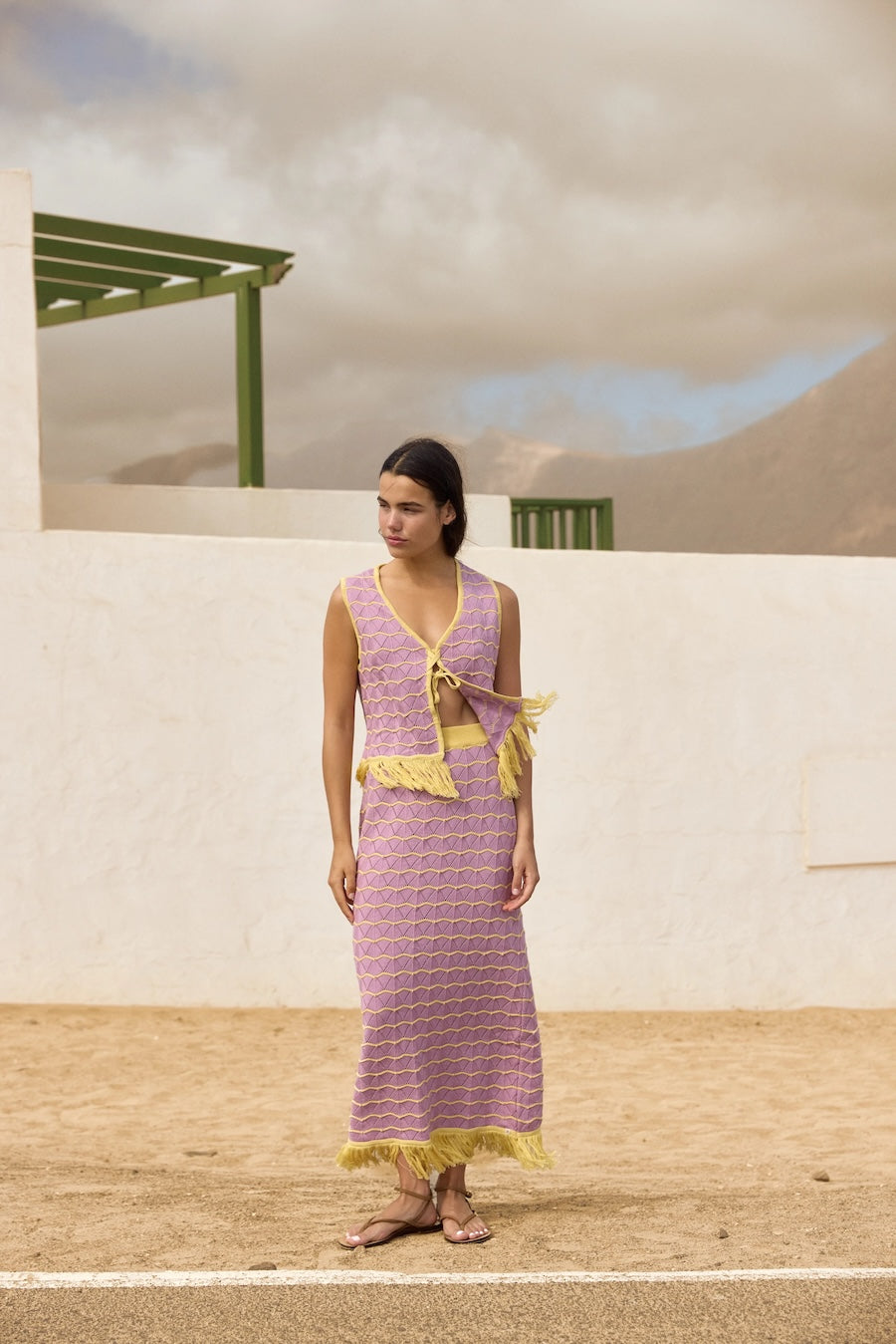 Woman wearing a pink skirt and top set with yellow accents standing in front of a white wall and green structure.