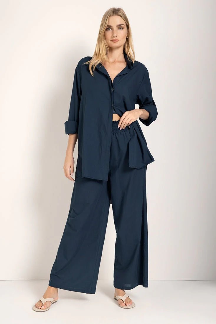 Woman wearing a button down shirt with wide leg pants. She's standing against a white background and looking at the viewer. The purpose of this image is to highlight the pants.