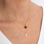 Close up image of a woman's neckline. She's wearing two necklaces. One has a charm of a Leo zodiac sign and the other has a Tiger's Eye stone. The purpose of this image is to highlight the necklaces, which are made in Colombia and sold in Studio City, at EVAMAIA boutique.