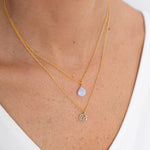 Close up image of a woman's neckline. She's wearing two necklaces. One has a charm of a Cancer zodiac sign and the other has a Blue Ribbon Agate stone. The purpose of this image is to highlight the necklaces, which are made in Colombia and sold in Studio City, at EVAMAIA boutique.