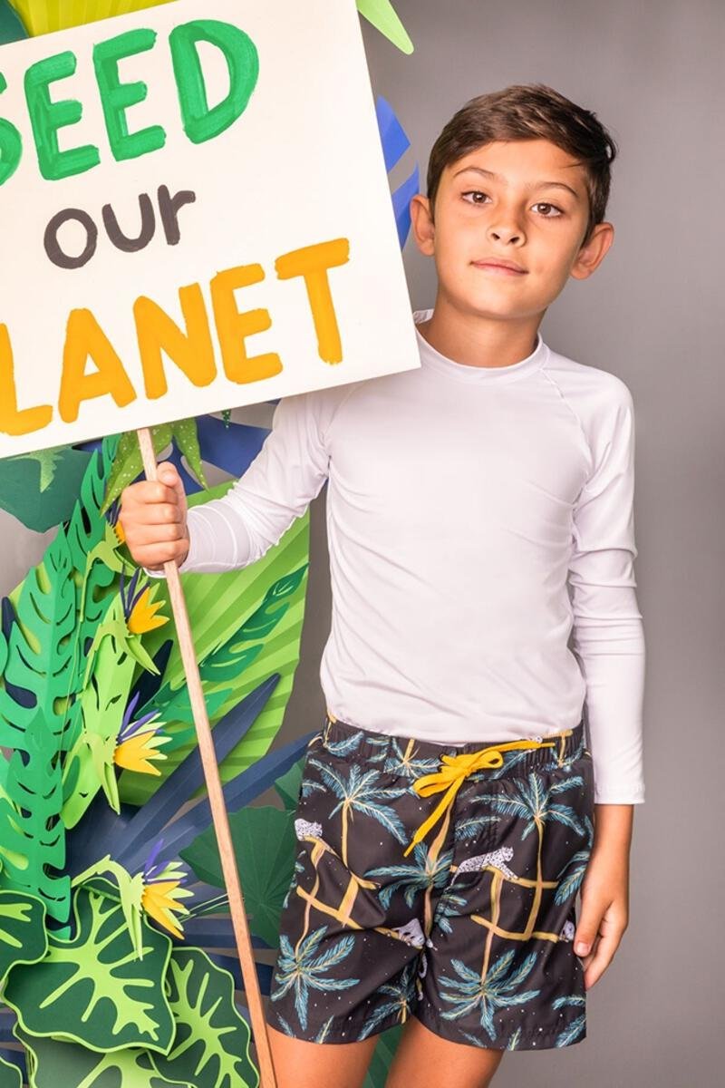 A boy wearing the A Dormir Swim Trunks for Kids. He is standing over a grey background with  leaf paper props. He is holding a sign that says "SEED OUR PLANET."