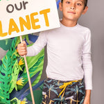 A boy wearing the A Dormir Swim Trunks for Kids. He is standing over a grey background with  leaf paper props. He is holding a sign that says "SEED OUR PLANET."