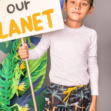 A boy wearing the A Dormir Swim Trunks for Kids. He is standing over a grey background with  leaf paper props. He is holding a sign that says "SEED OUR PLANET."