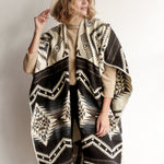Woman modeling front of Arizona Poncho. Arizona Poncho features an overall brown themed geometric pattern all over. 
