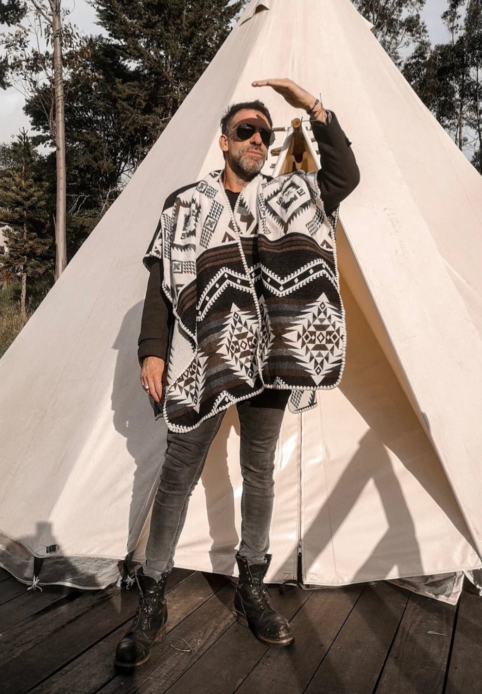 Man wearing the Arizona Poncho with Hood. He is standing on a wood floor, in front of a white tent in a natural setting. He is looking away into the distance.