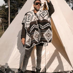 Man wearing the Arizona Poncho with Hood. He is standing on a wood floor, in front of a white tent in a natural setting. He is looking away into the distance.