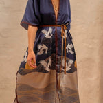 Model wearing the Asia Kimono. This close up image includes the blue kimono and the details of sand plains and birds, a setting sun, all paired with a gold necklace. She is standing barefoot on rug next to a large mirror and limewash walls behind her.