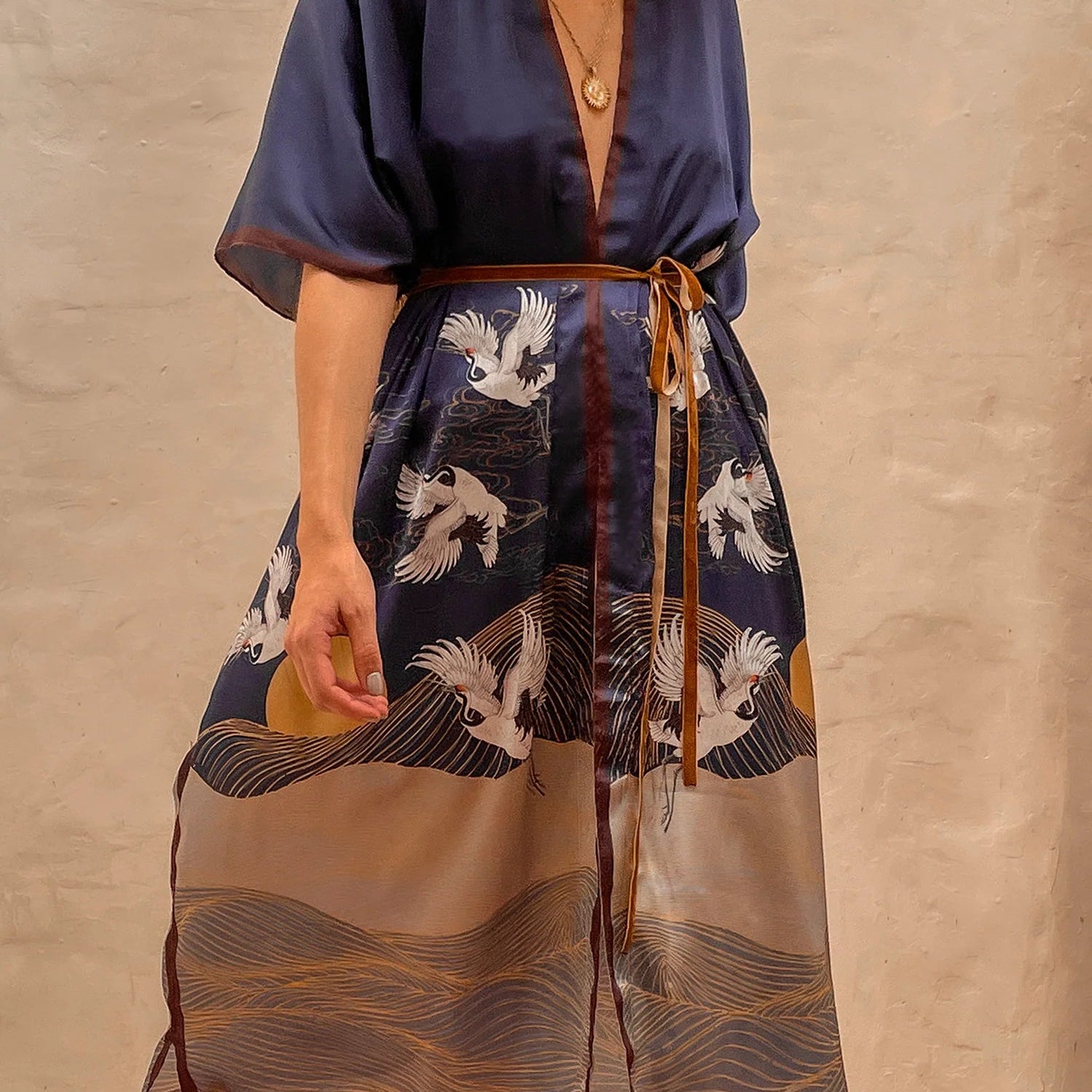 Model wearing the Asia Kimono. This close up image includes the blue kimono and the details of sand plains and birds, a setting sun, all paired with a gold necklace. She is standing barefoot on rug next to a large mirror and limewash walls behind her.