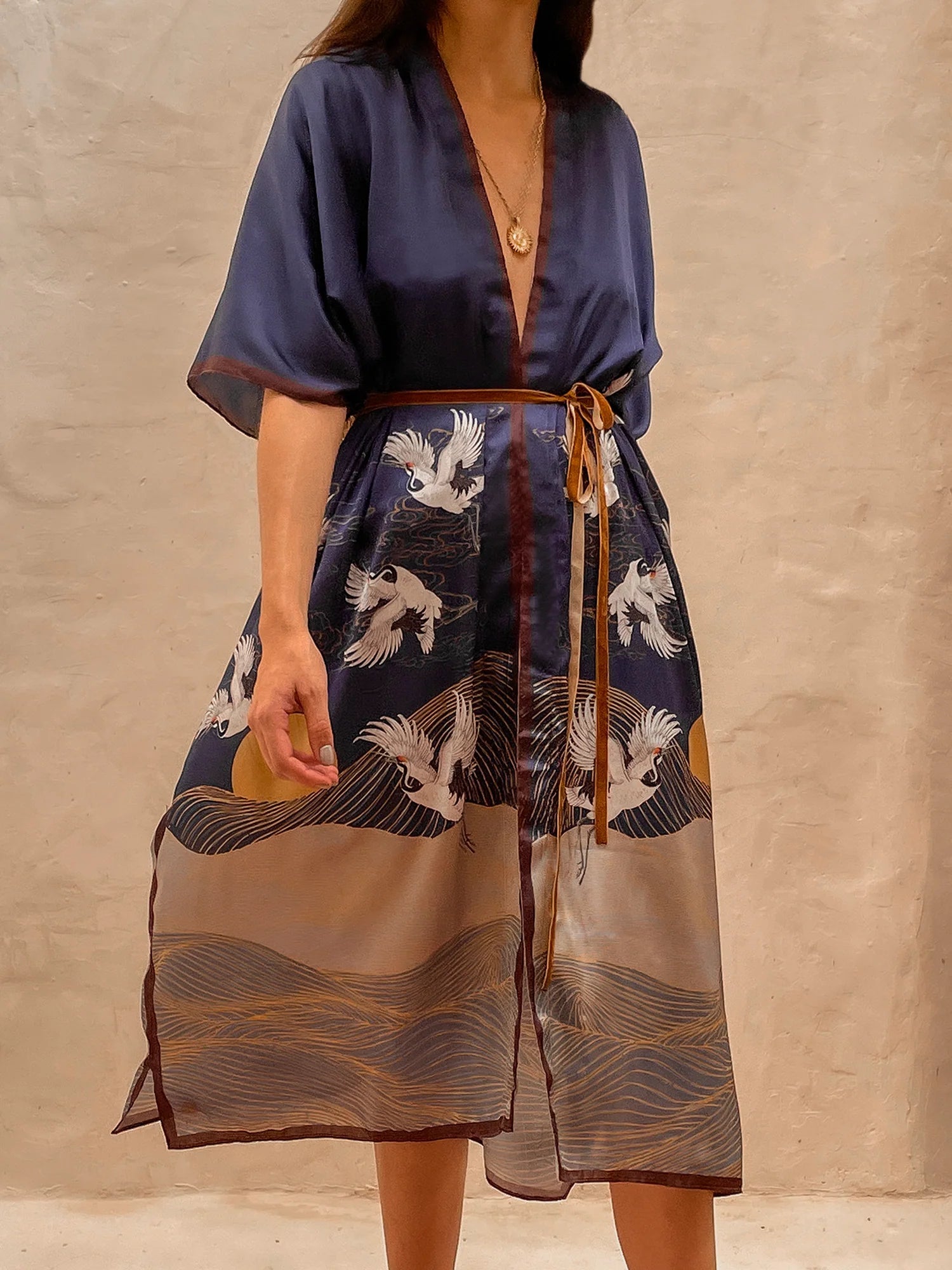 Model wearing the Asia Kimono. This close up image includes the blue kimono and the details of sand plains and birds, a setting sun, all paired with a gold necklace. She is standing barefoot on rug next to a large mirror and limewash walls behind her.