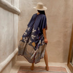 Back of the flowy Asia Kimono. The model paired this kimono with a cream colored hat and gold jewelry. She is standing barefoot on rug next to a large mirror and limewash walls.