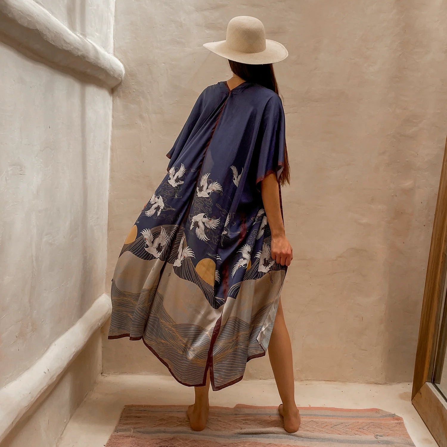 Back of the flowy Asia Kimono. The model paired this kimono with a cream colored hat and gold jewelry. She is standing barefoot on rug next to a large mirror and limewash walls.