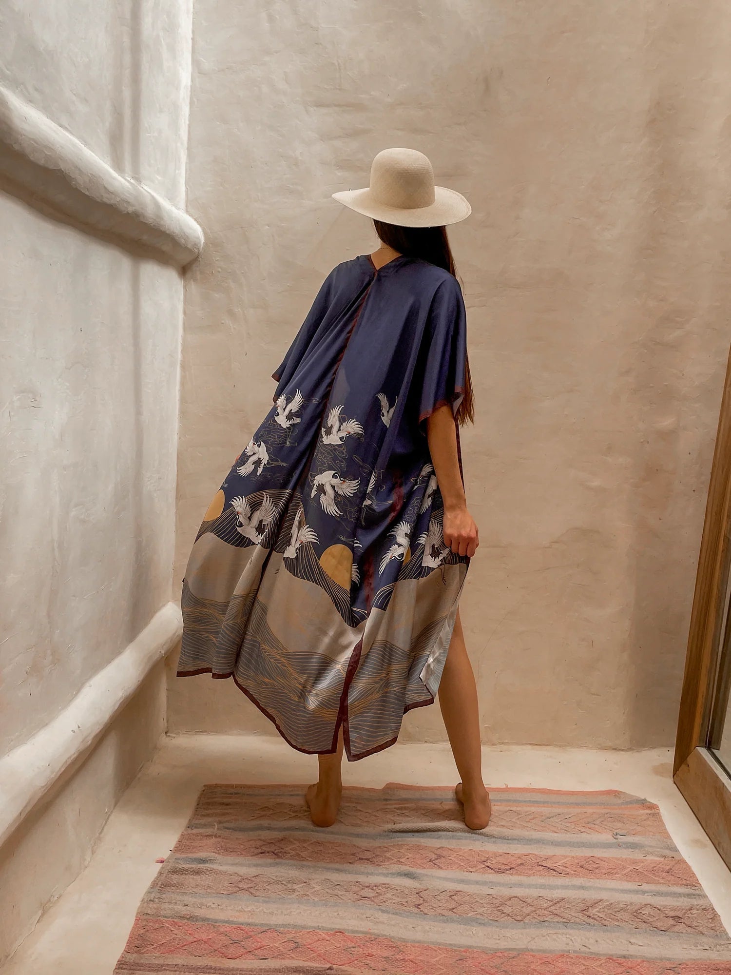 Back of the flowy Asia Kimono. The model paired this kimono with a cream colored hat and gold jewelry. She is standing barefoot on rug next to a large mirror and limewash walls.