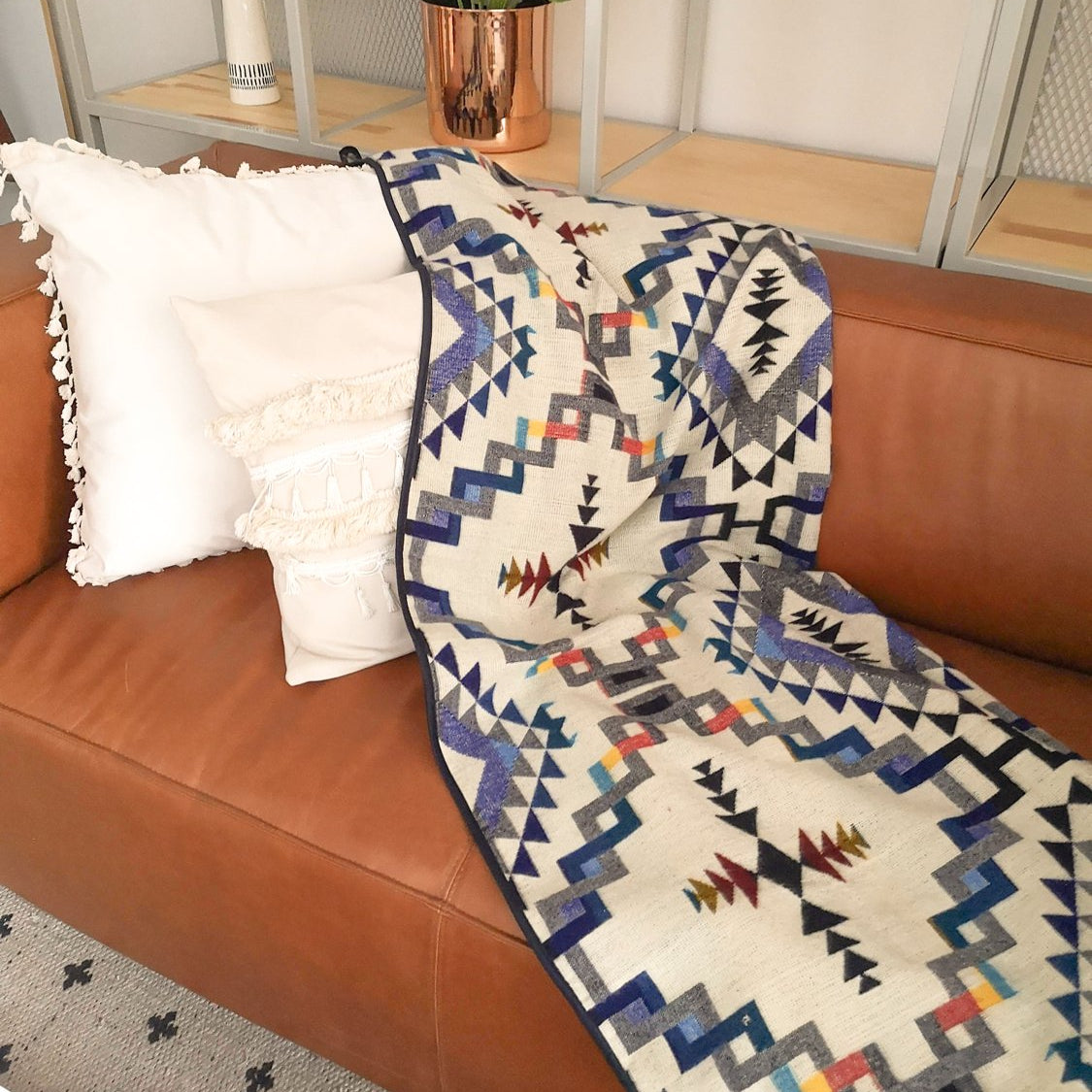 Cream geometric blanket. The pattern is a blend of colors like navy blue ,grey, black, yellow, etc. The blanket is draped over a brown leather couch styled with two white pillows.