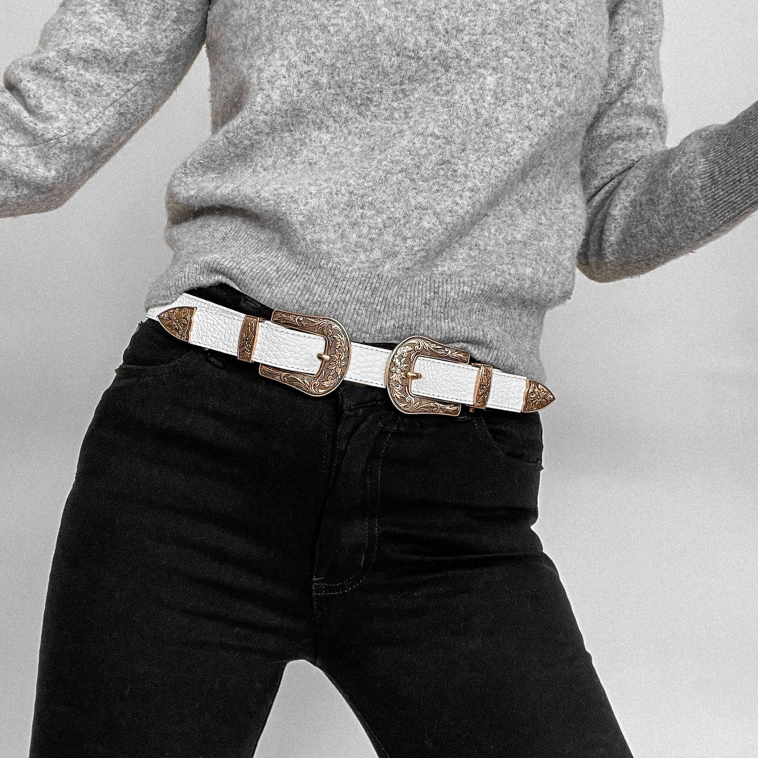 Model is wearing the White Florentino Leather Belt by Plissé. This belt features two buckles with a Western floral detail. Made of 100% Leather. Belt is styled with a grey long sleeve top and black jeans.