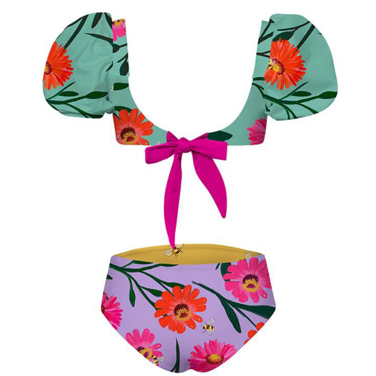 Back of cute two piece swimsuit for girls. The top has a puffy sleeve with a flower and bee pattern and bow tie in the back for secure wear. The bottom is reversible with the same pattern. The suit is displayed over a white background.