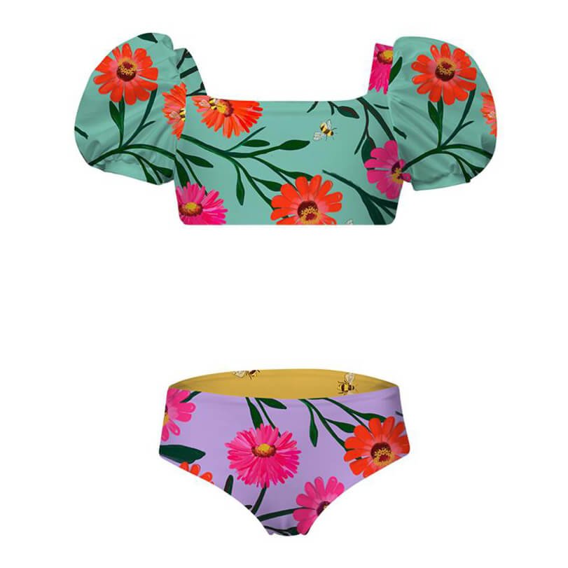 Front of cute two piece swimsuit for girls. The top has a puffy sleeve with a flower and bee pattern. The bottom is reversible with the same pattern. The suit is displayed over a white background.