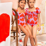 Two girls posing against a chair in an art studio wearing different versions of the Bubble One Piece in the heart pattern. 