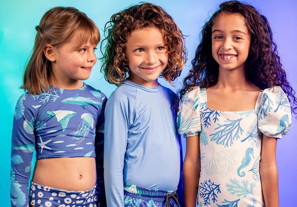 3 kids over a similar gradient background wearing different swimsuits. One of the little girls is wearing the Fiesta En El Mar two piece bikini. 