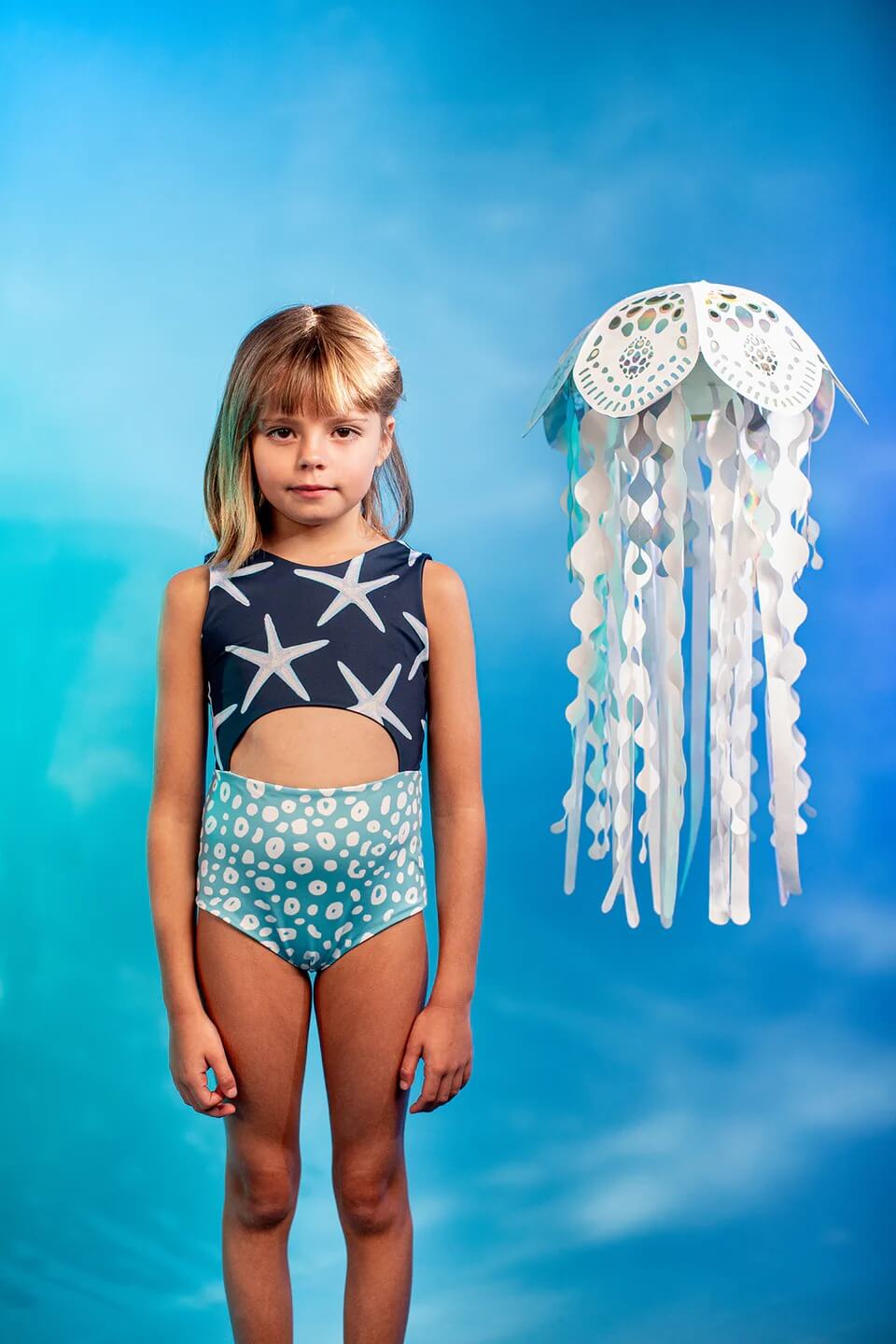 Girls' Sea Star Trikini in Deep Blue worn by little girl. Gradient background and cute white jellyfish paper prop up in the air.