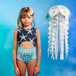 Girls' Sea Star Trikini in Deep Blue worn by little girl. Gradient background and cute white jellyfish paper prop up in the air.