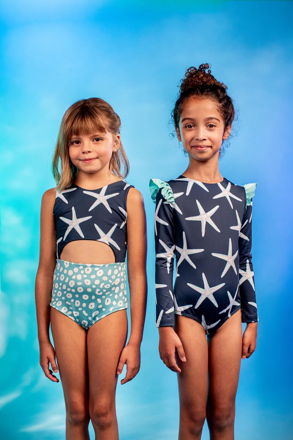 Girls' Sea Star Trikini in Deep Blue worn by little girl. She is posing in front of blue gradient background with another girl in another one piece.