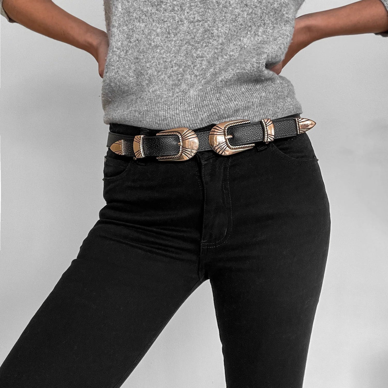 Model wearing the Insignia Belt. This belt features two buckles with a Western gold detail. Made of 100% Leather. Belt is styled with a grey long sleeve top and black jeans.