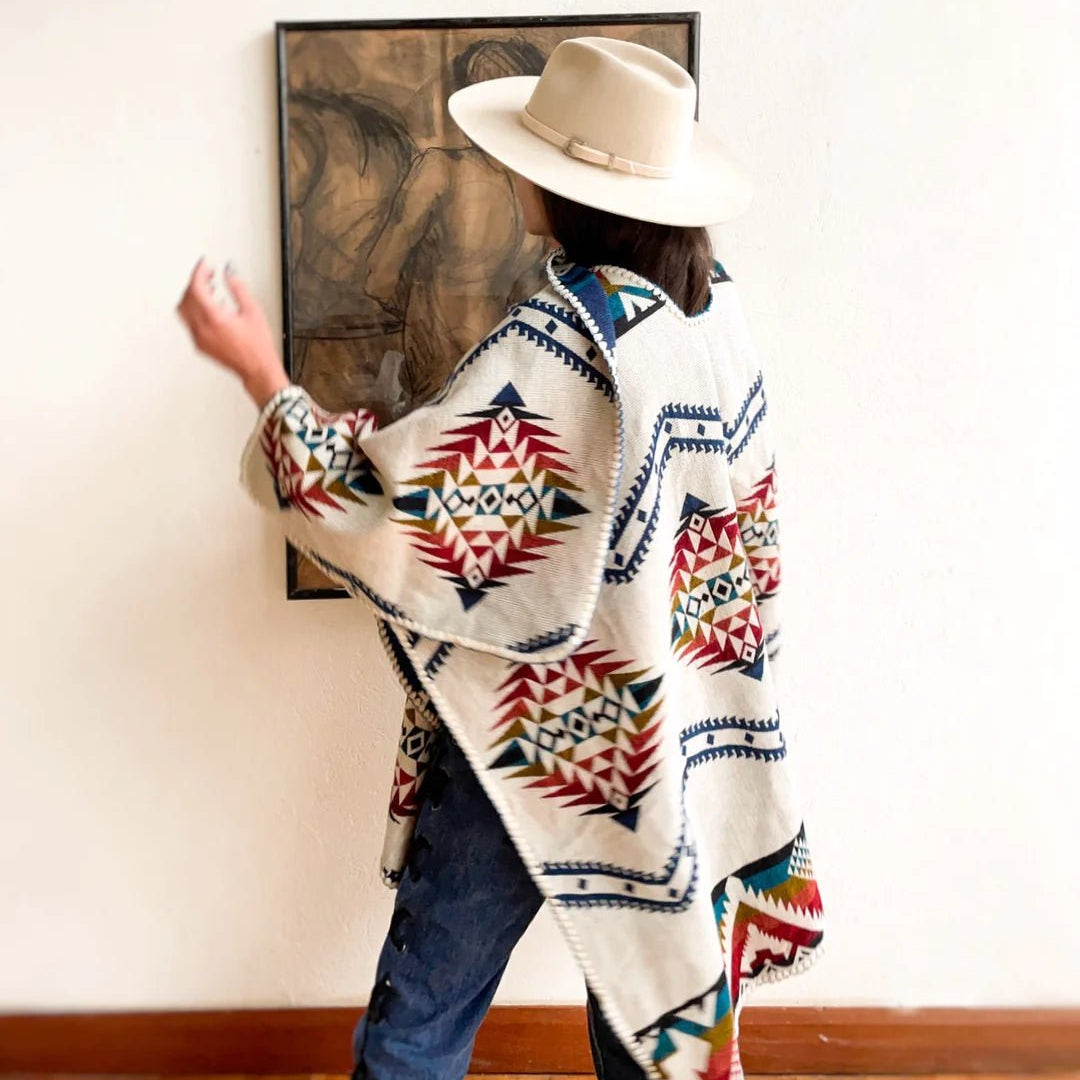 Model wearing and showing the back of the Jalisco Poncho. It is layered over blue jeans and paired with a white hat. Model is standing near white wall with hardwood floors and a small painting on wall.