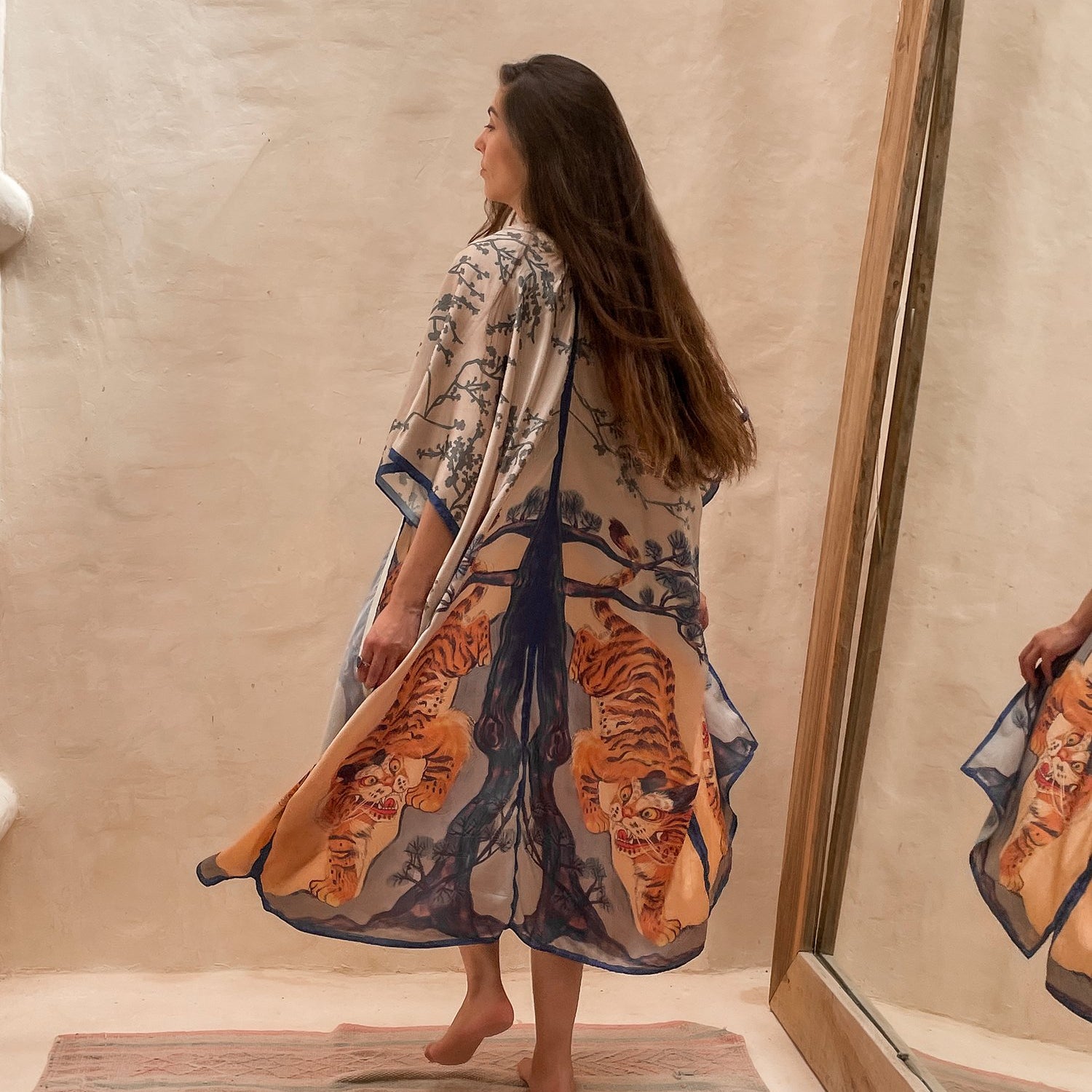 Model wearing the Kimono Bengal Tiger. She is towards the wall with a large mirror next to her. The design features a mirrored design of a large tree, nature details, and the bengal tiger.